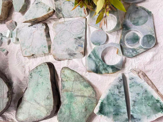 An overhead view of multiple jadeite rough stones on a white surface. Some of the stones are already cut and have chalk circles drawn on them, marking out shapes for jade bangles and pendants.