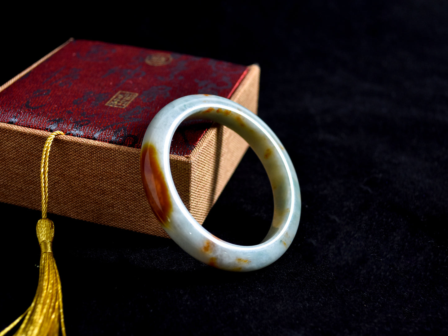 White jade ring with brown inclusions on a dark surface with a red box and yellow tassel in the background.