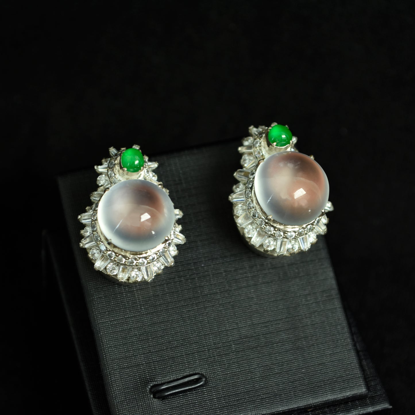 A pair of imperial green jadeite and translucent cabochon earrings with diamond pavé, elegantly displayed on black silk.