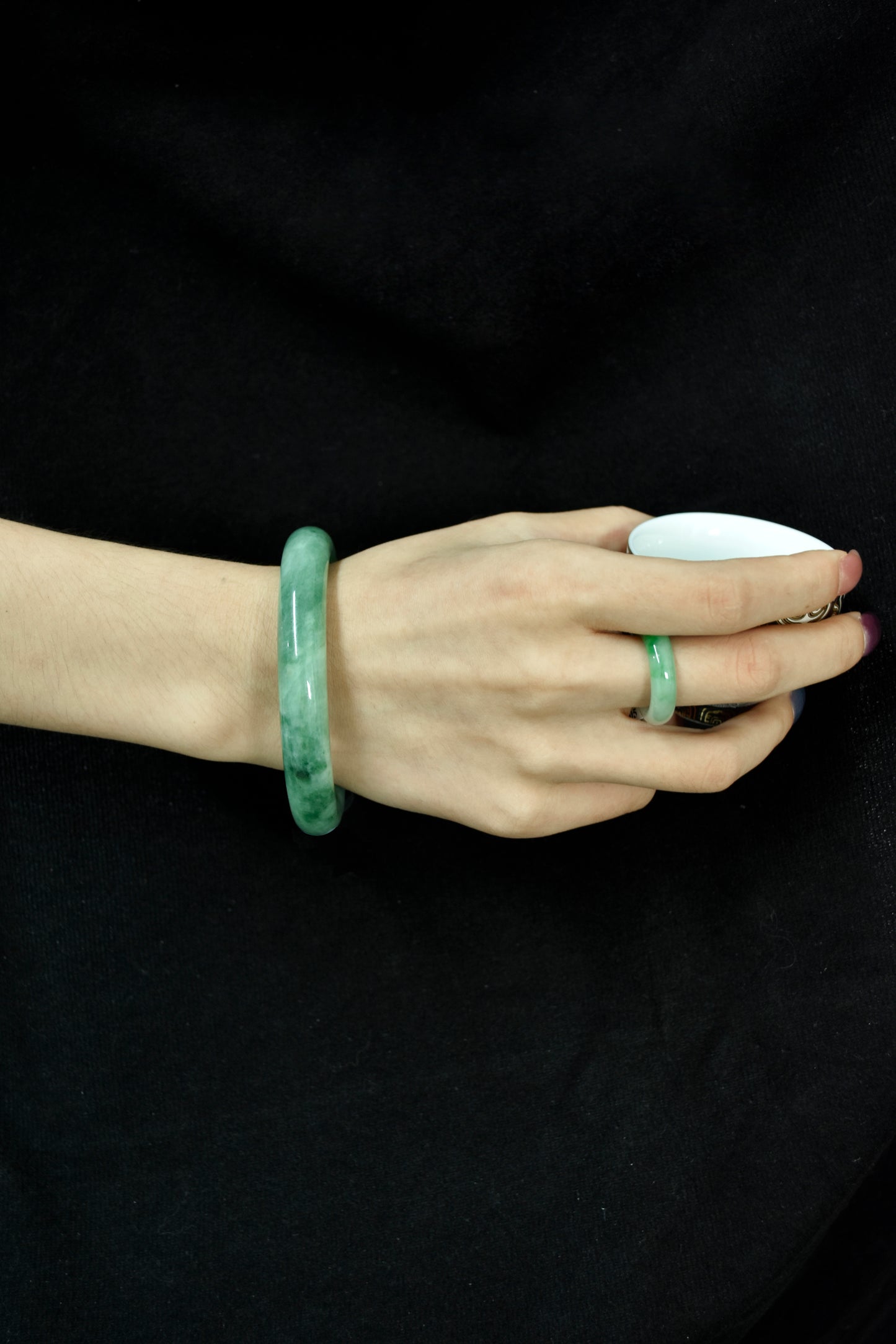 Model wearing a vivid green jadeite bangle and matching jadeite ring on hand, set against a black fabric background.