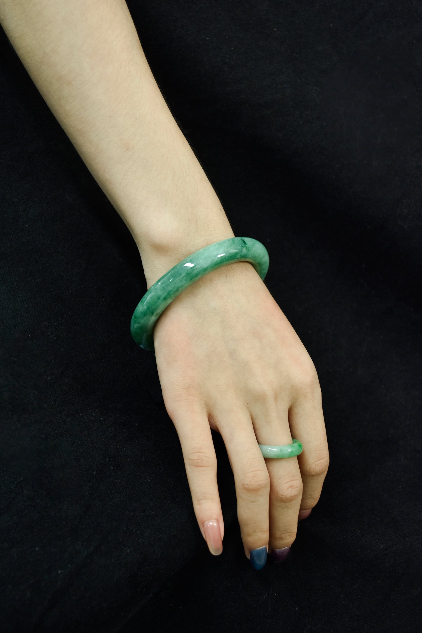 Model wearing a vivid green jadeite bangle and matching jadeite ring on hand, set against a black fabric background.