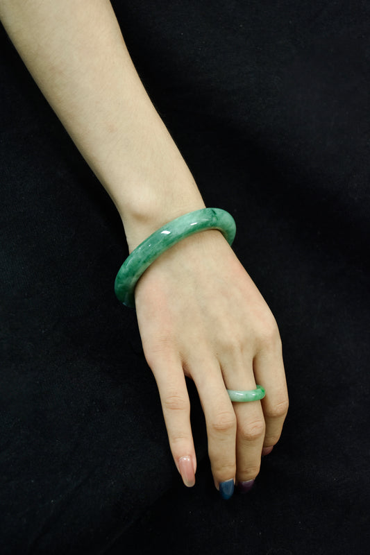 Model wearing a vivid green jadeite bangle and matching jadeite ring on hand, set against a black fabric background.