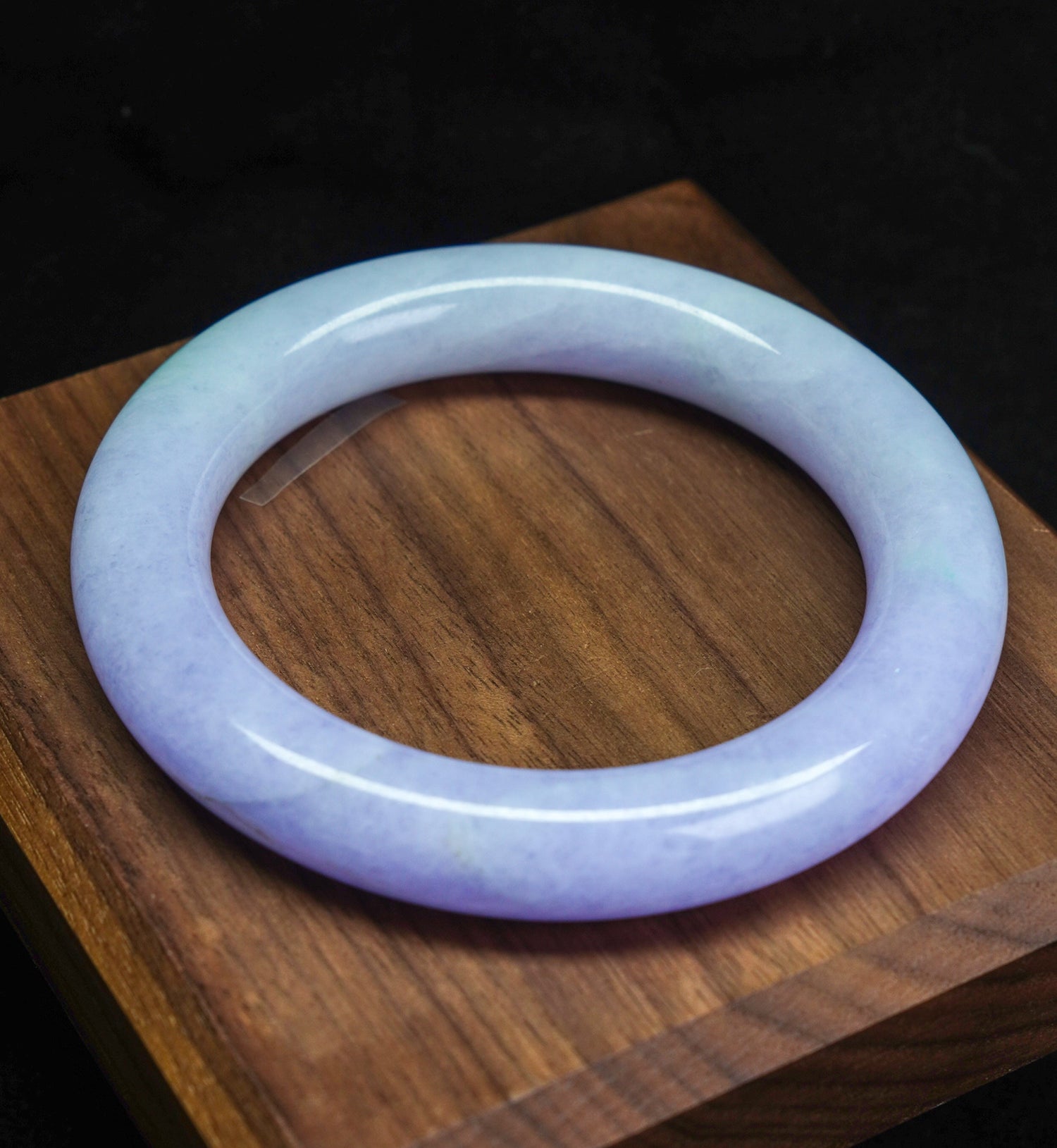 Lavender jadeite bangle with a smooth round shape, resting on a wooden display. High-grade jade from TATHATĀ.