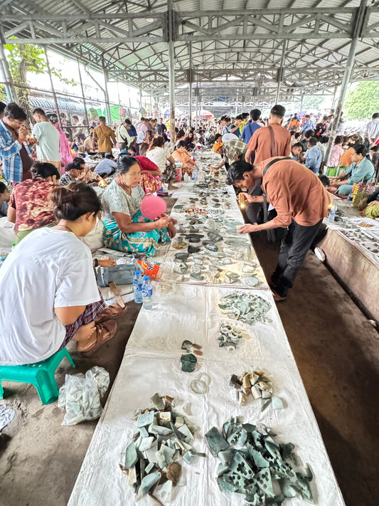 A bustling open-air market in Myanmar with people examining various rough and polished jadeite pieces laid out on white cloths on the ground.