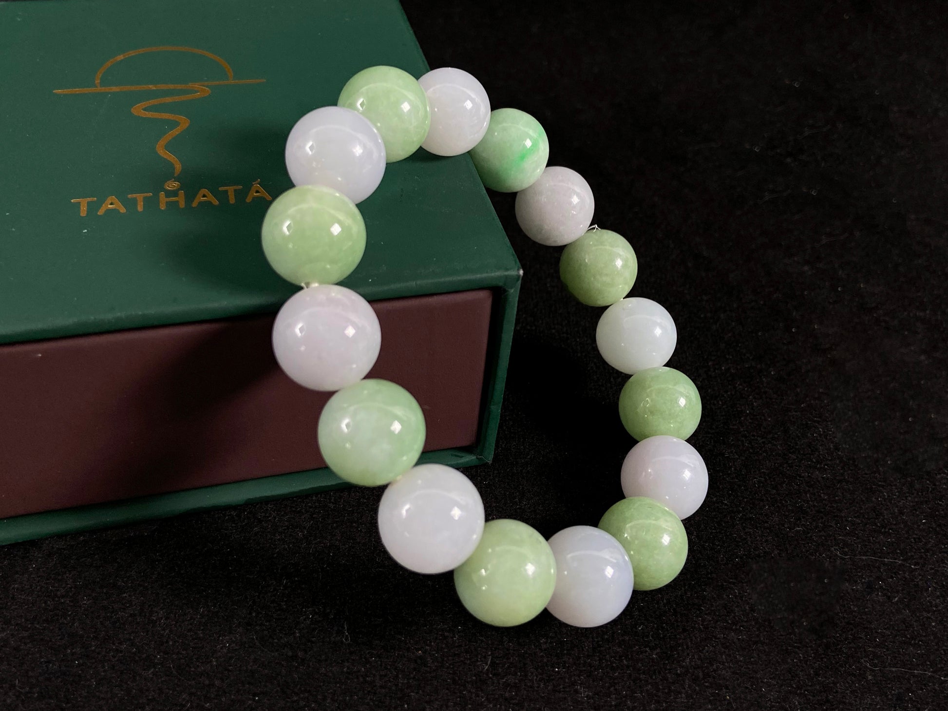 Beaded bracelet with green and white beads on a dark background, with Tathata brand box in the background.