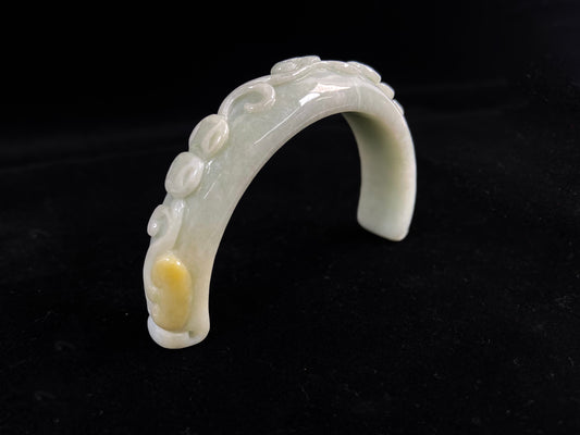 This piece features a creamy white jade half-bangle carved with coins, a Ruyi scepter, and ganoderma, accented with sprinkled gold.