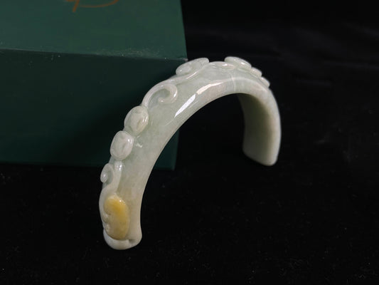 This piece features a creamy white jade half-bangle carved with coins, a Ruyi scepter, and ganoderma, accented with sprinkled gold.