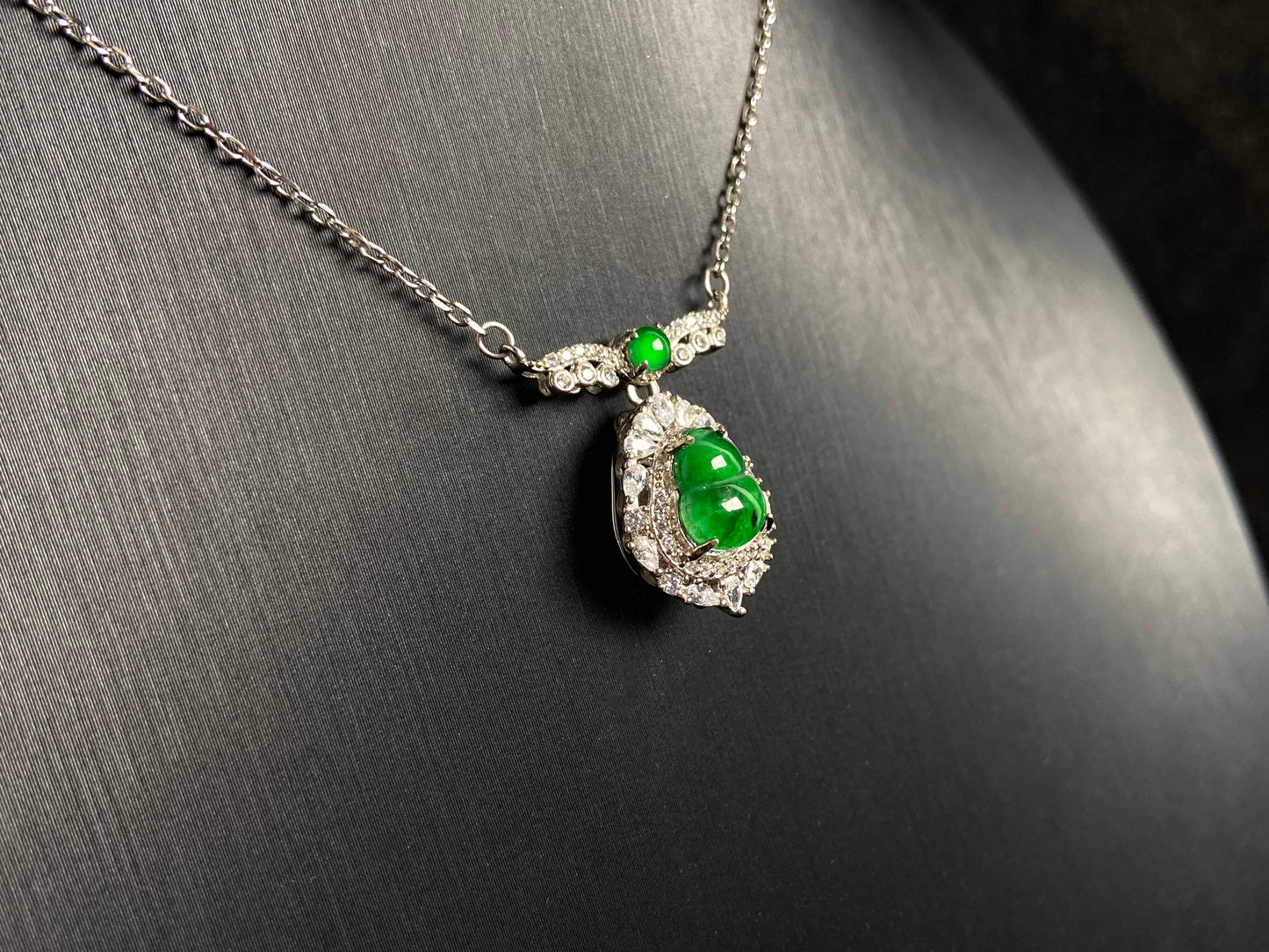 A silver framed green jadeite Gourd pendant, topped with a bead and backed by a gold money bag symbolizing wealth and power.