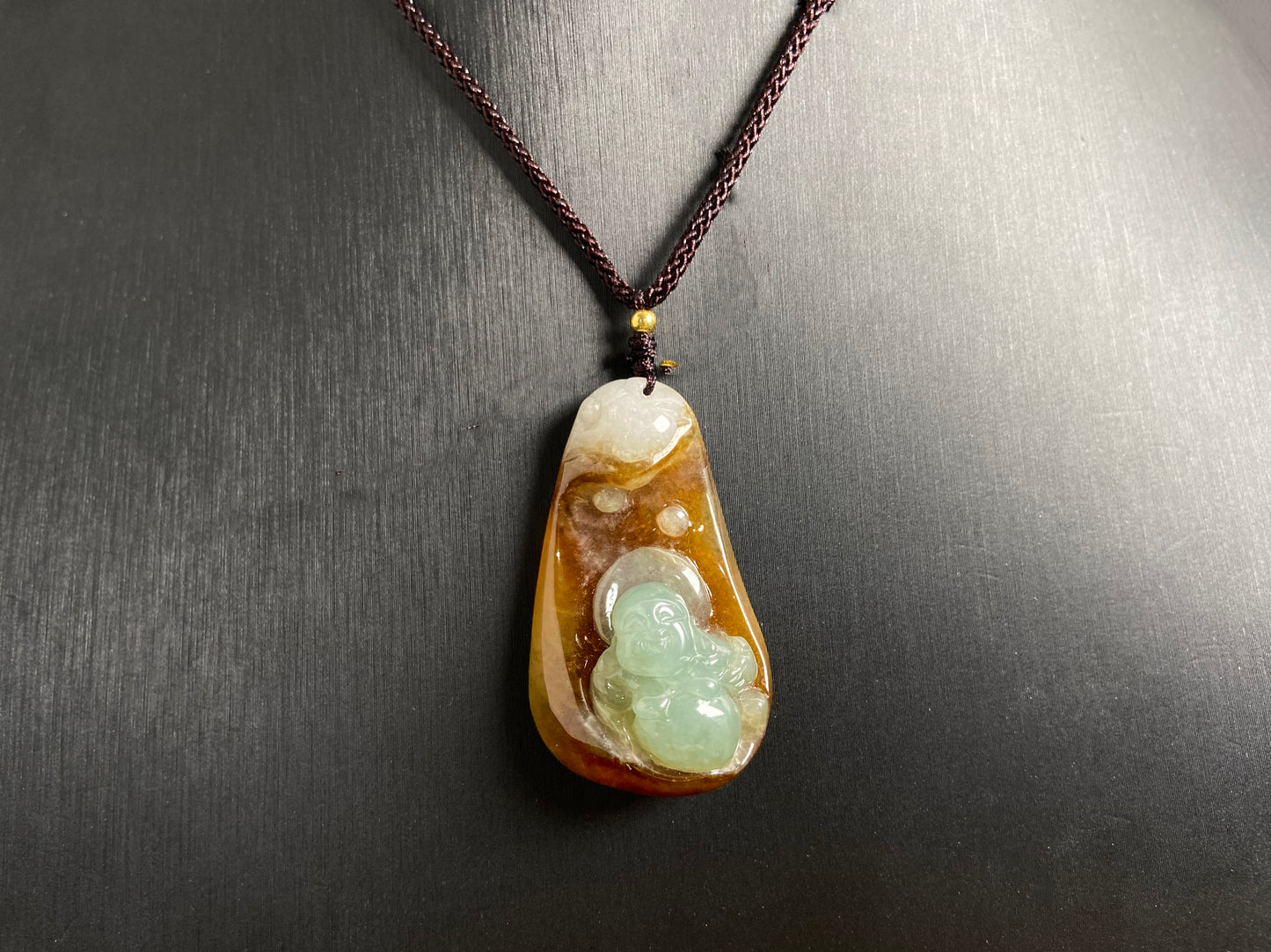 This jade pendant features a brown jade base with white accents, cradling a light green reclining Maitreya Buddha in its design.