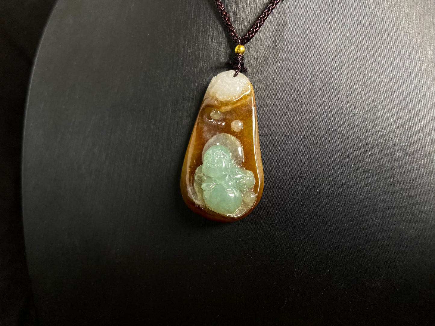 This jade pendant features a brown jade base with white accents, cradling a light green reclining Maitreya Buddha in its design.