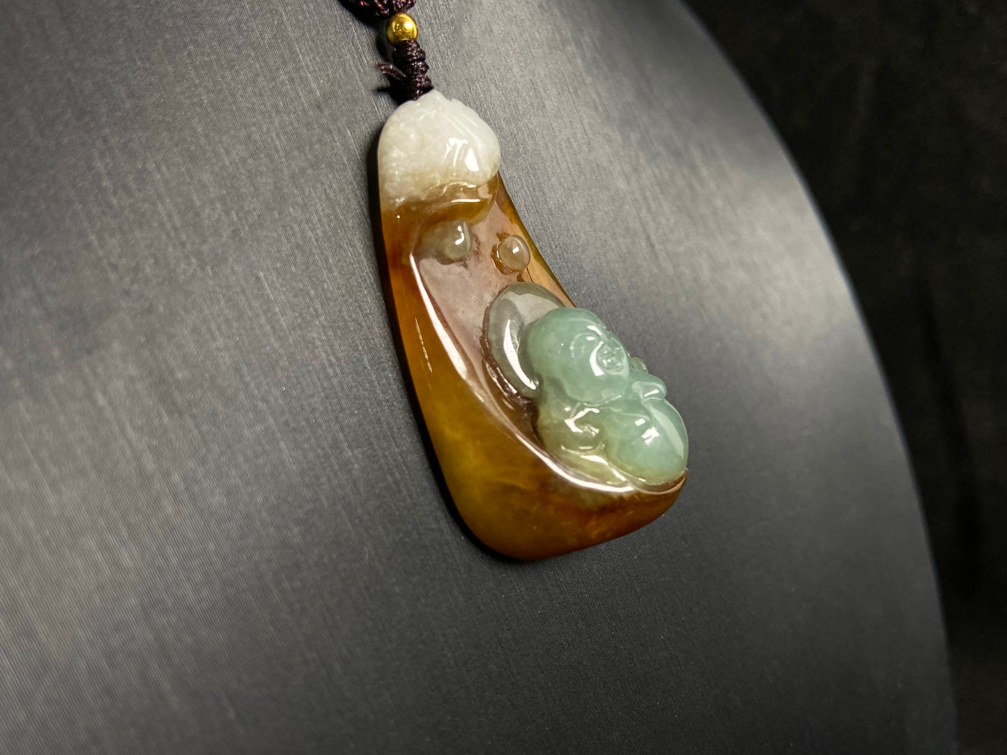 This jade pendant features a brown jade base with white accents, cradling a light green reclining Maitreya Buddha in its design.