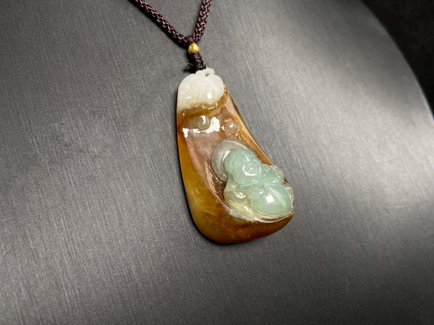 This jade pendant features a brown jade base with white accents, cradling a light green reclining Maitreya Buddha in its design.