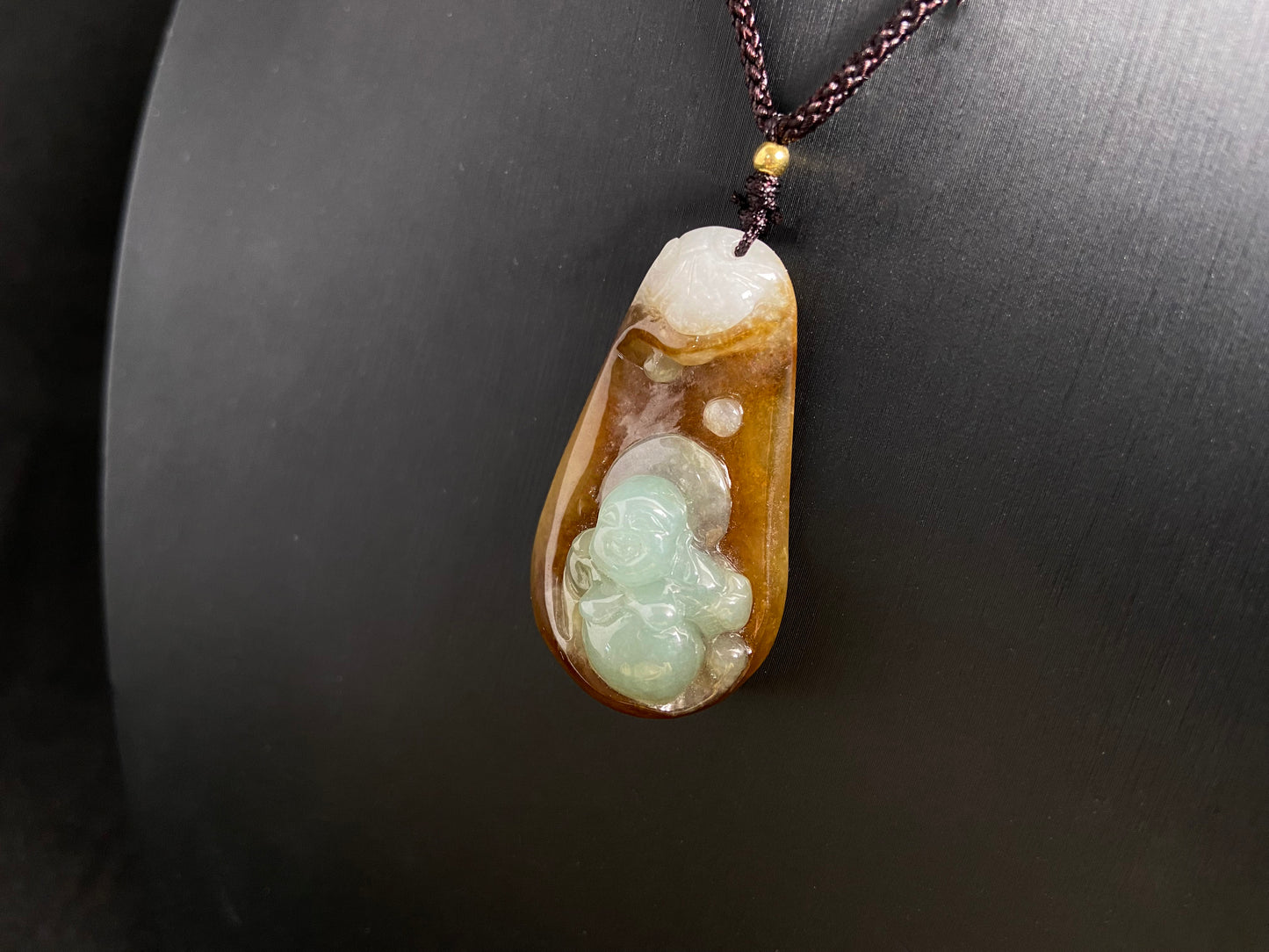 This jade pendant features a brown jade base with white accents, cradling a light green reclining Maitreya Buddha in its design.