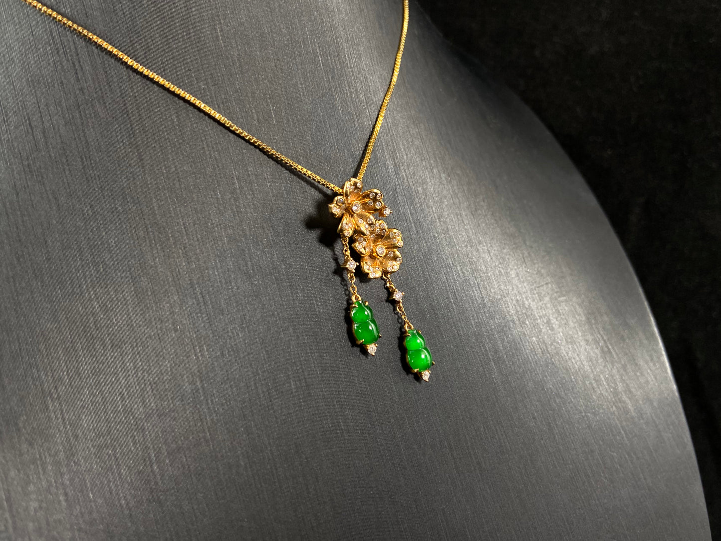 This elegant pendant showcases two emerald green jade gourds suspended from delicately crafted 18k gold flowers.