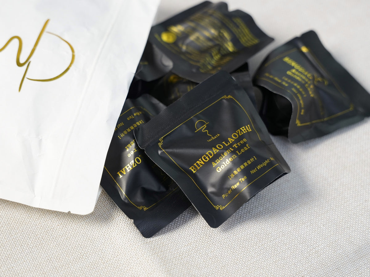 TATHATA Bingdao Laozhai Ancient Tree Golden Leaf raw Pu‑er tea, single serve black sachet packets spilled out from white pouch, premium Yunnan raw puer tea.