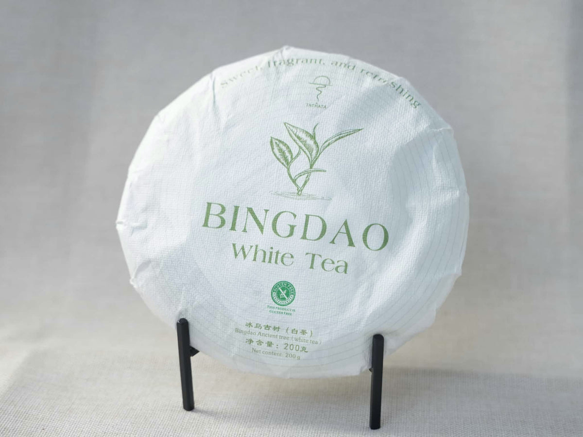 A round cake of aged Icelandic ancient-tree white tea (200g Yunnan Bingdao white tea), wrapped in patterned paper, displayed on a small black stand against a soft linen background.