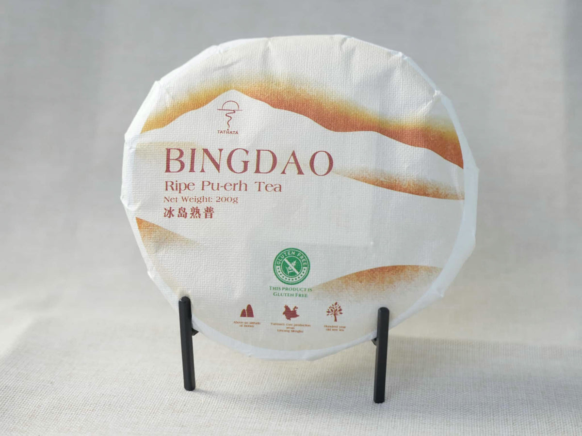 This 200g Bingdao ripe Pu-erh tea cake features artistic landscape-themed packaging and is displayed on a small black stand.