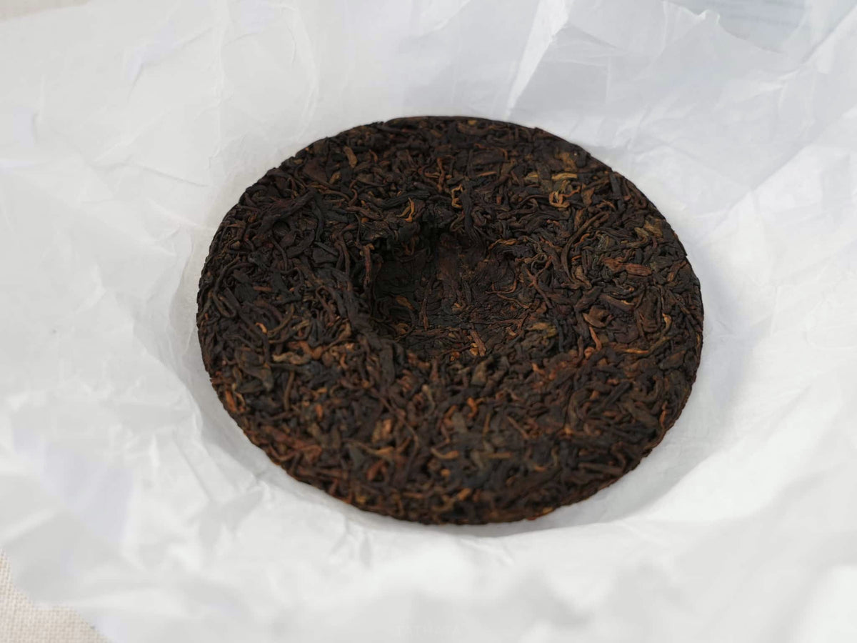 A close-up shot of a Bingdao ripe Pu-erh tea cake: the dark brown fermented tea leaves rest on white wrapping paper, clearly revealing the details of the pressed tea cake's surface.