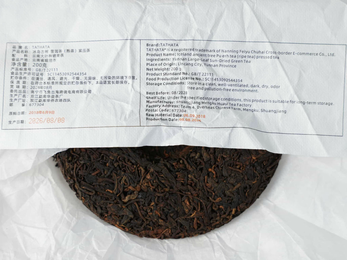 A close-up of the “TATHATA” Iceland ancient-tree ripe Pu-erh tea cake. A label featuring product information in both Chinese and English is affixed to the wrapping paper, with a portion of the compressed tea cake visible beneath.