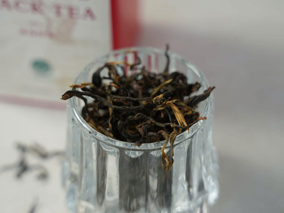 A close-up shot of premium Yunnan Bingdao black tea: curled loose-leaf tea in a clear glass, with blurred product packaging in the background.