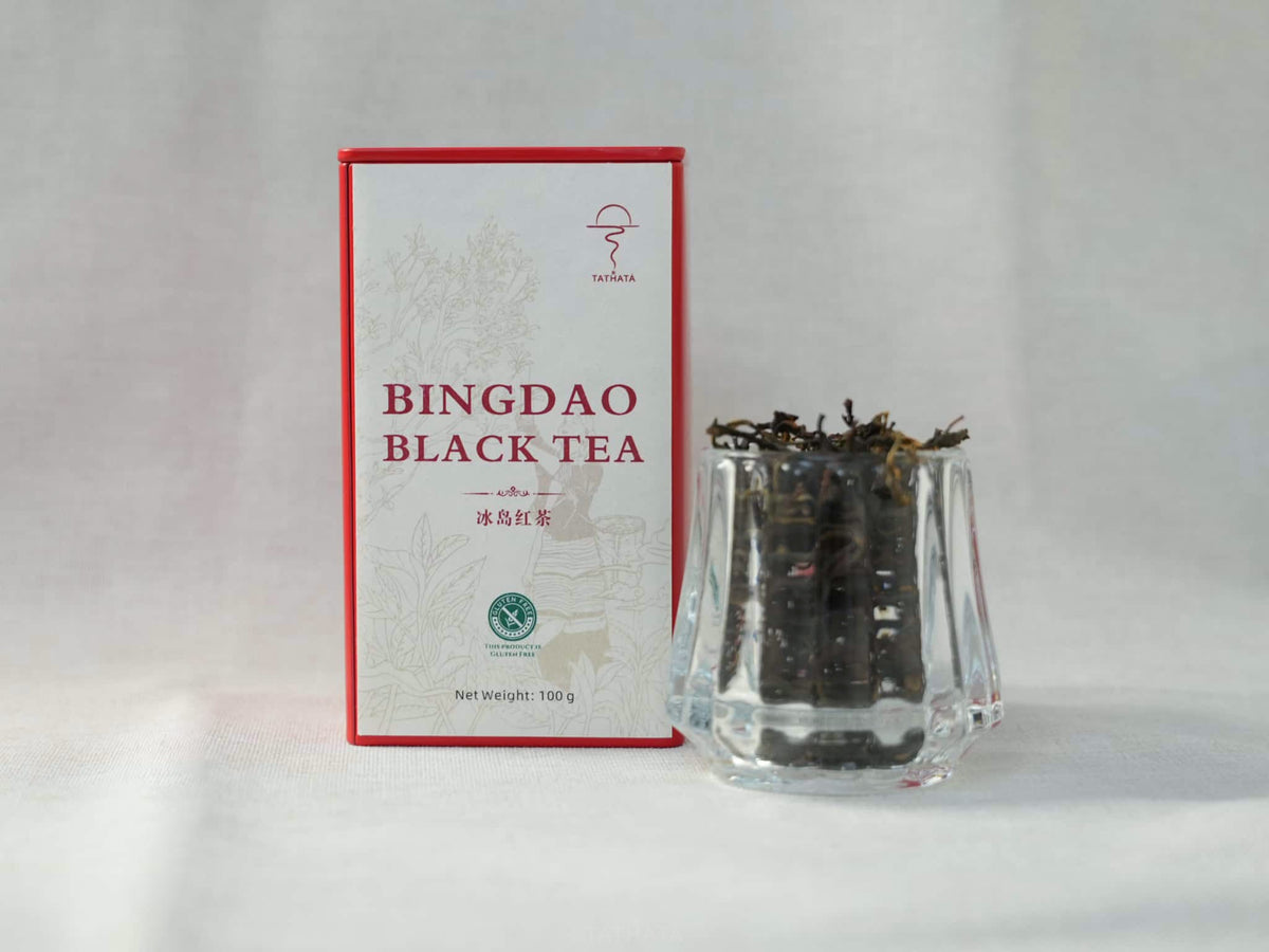 On a linen background, a clear glass cup containing loose-leaf tea sits next to a red TATHATA Icelandic black tea tin.