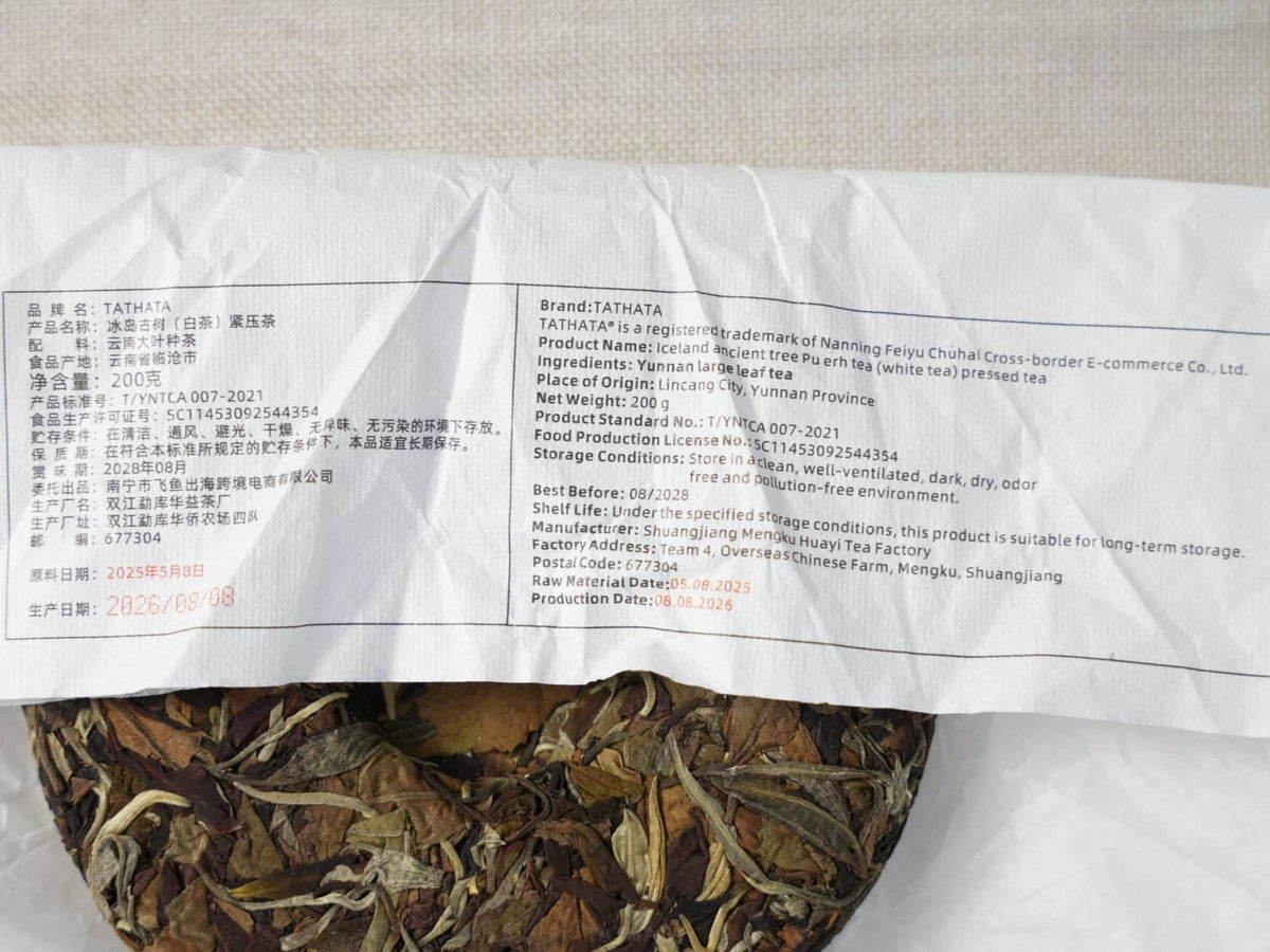 A close-up of a TATHATA compressed white tea cake made from ancient Iceland tea trees. The wrapping paper displays product specifications in both Chinese and English, with a portion of the tea cake visible beneath.