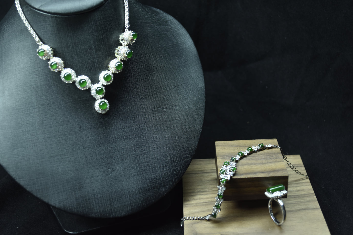 Silver necklace with green gemstones on a mannequin bust and wooden stand against a dark background.