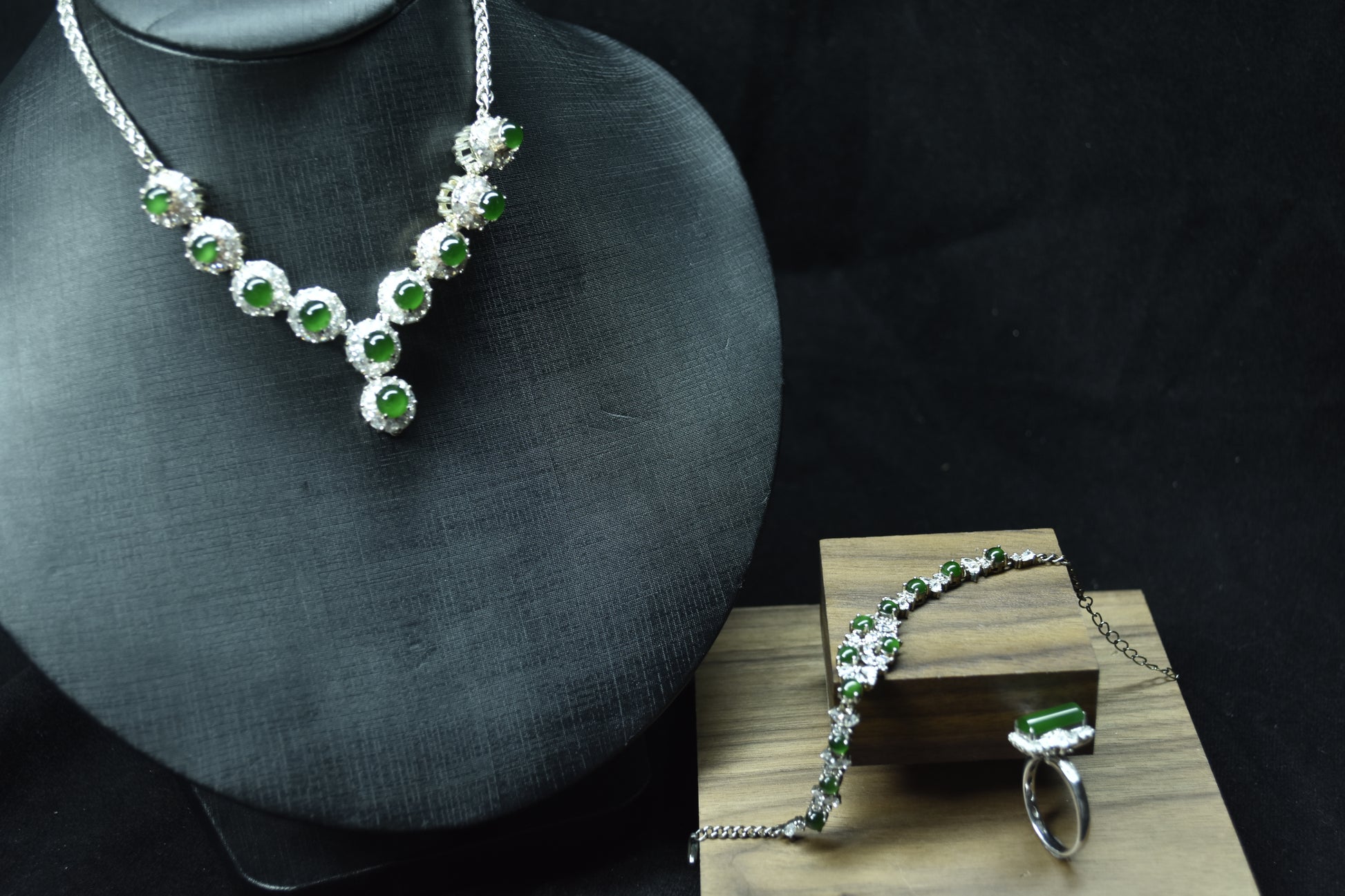 Silver necklace with green gemstones on a mannequin bust and wooden stand against a dark background.