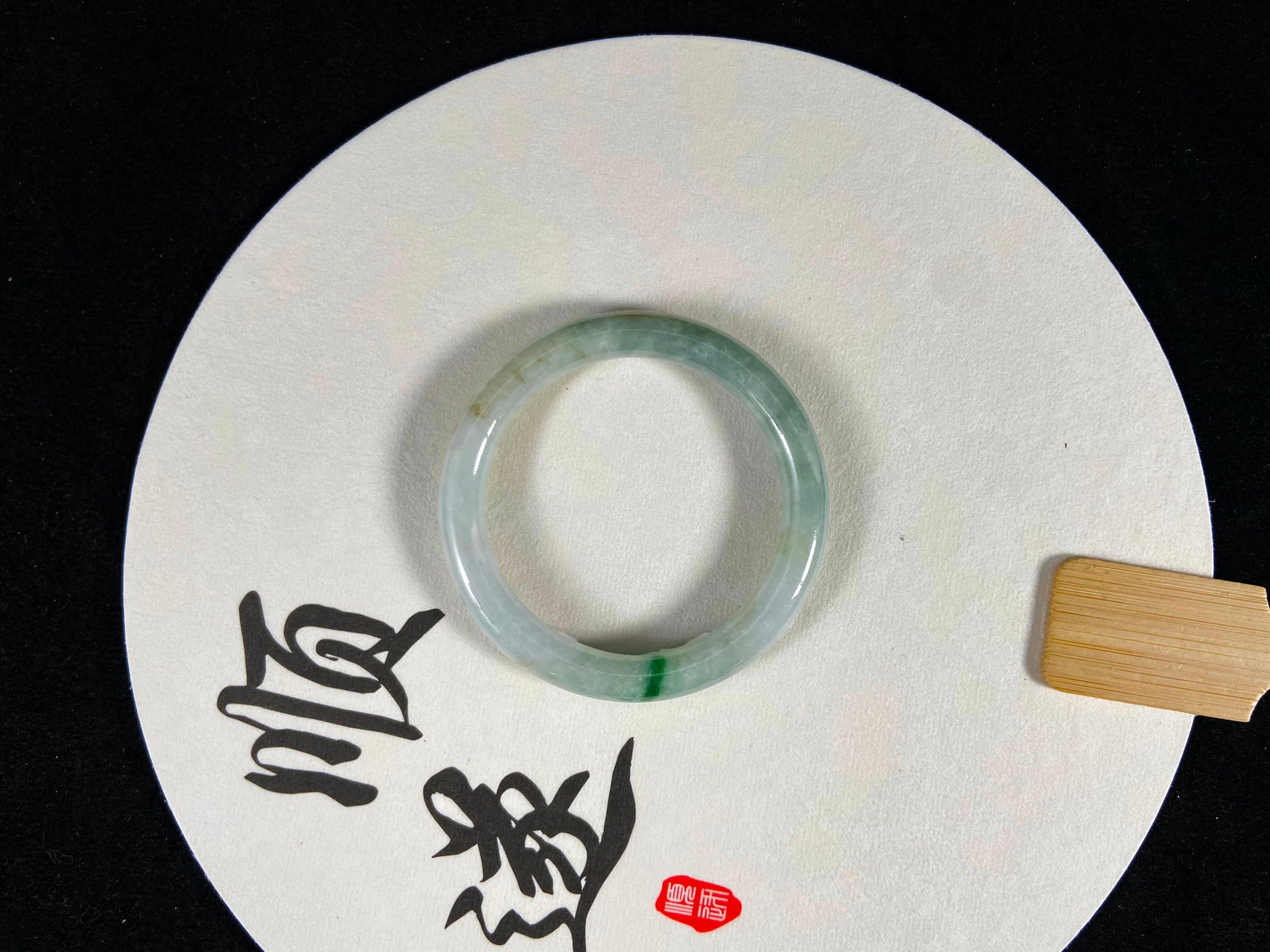A jade bangle with an emerald vein rests on a white fan inscribed with "Shun Sui".