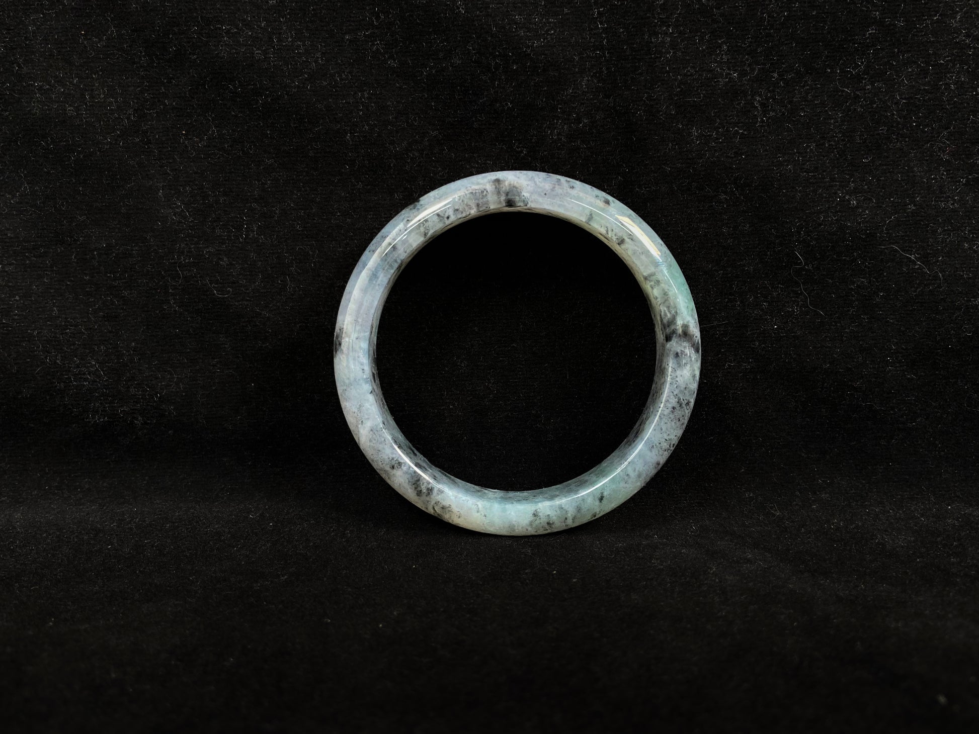 This jadeite bangle features a semi-transparent light green base with evenly distributed charcoal grey inclusions.
