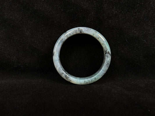 This jadeite bangle features a semi-transparent light green base with evenly distributed charcoal grey inclusions.