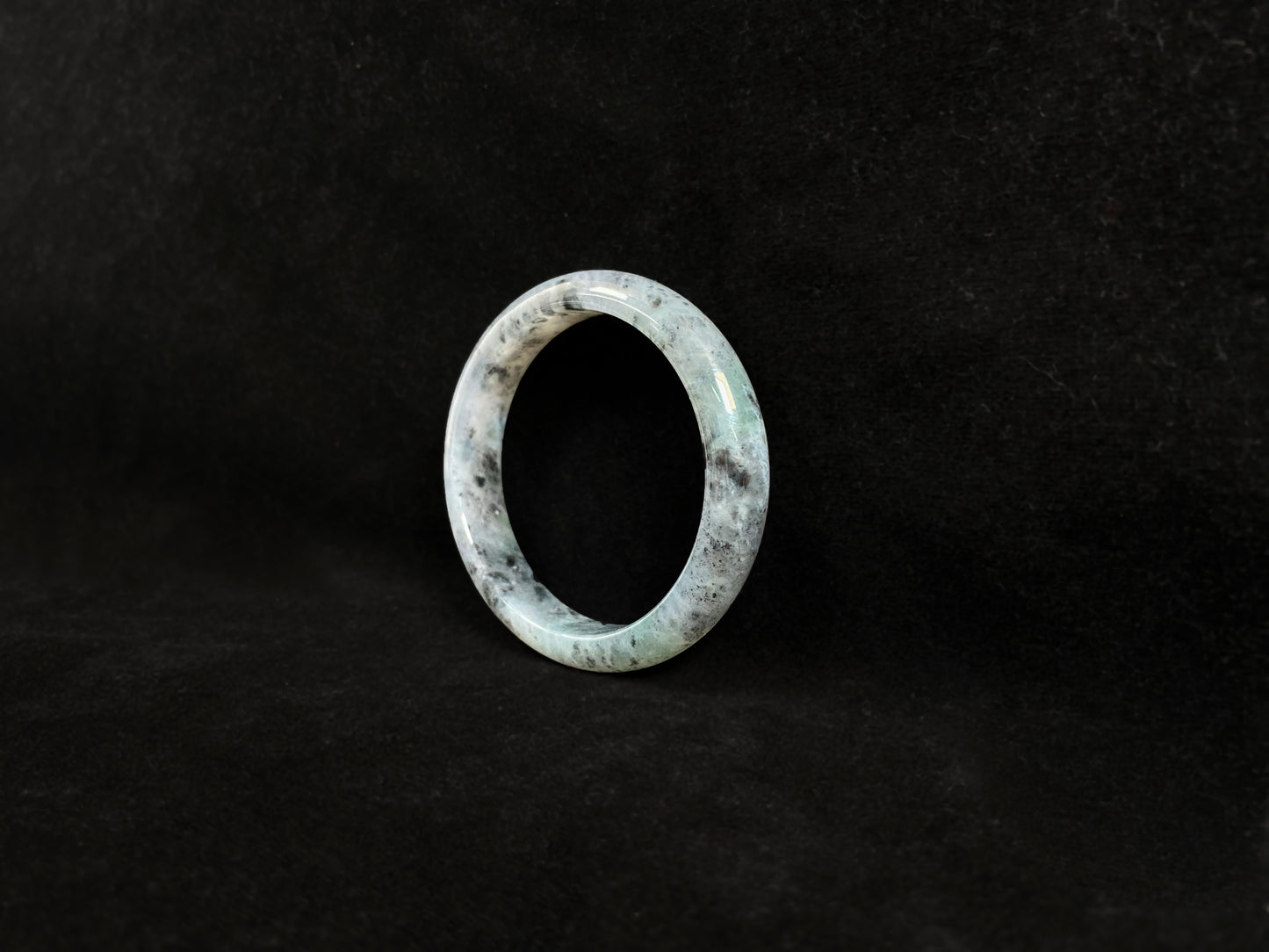 This jadeite bangle features a semi-transparent light green base with evenly distributed charcoal grey inclusions.