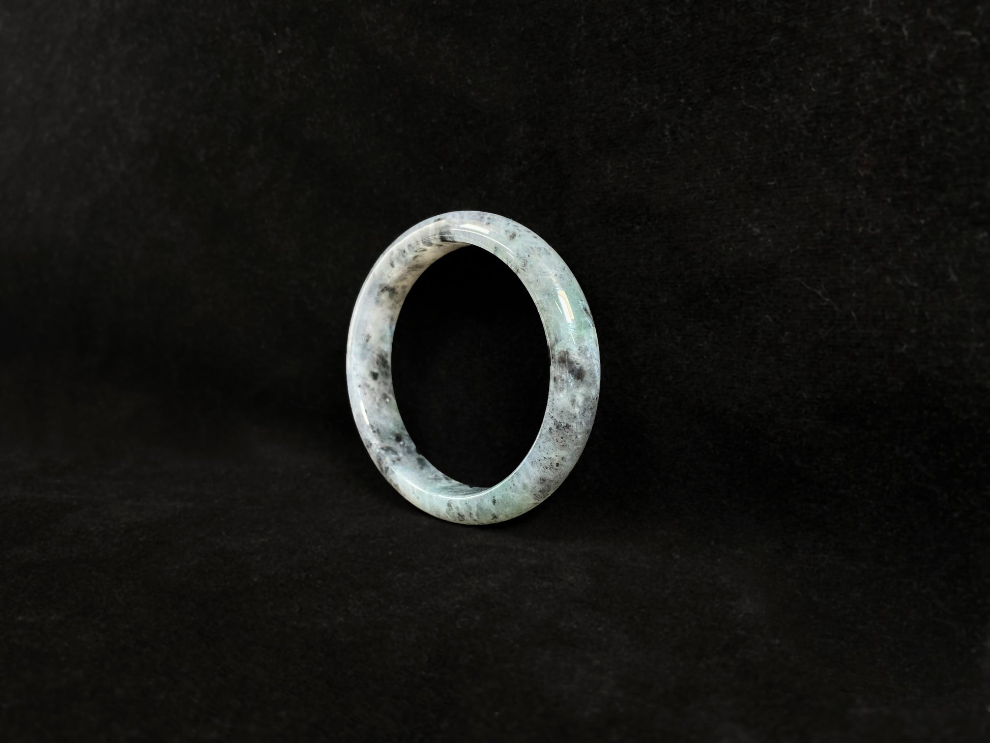 This jadeite bangle features a semi-transparent light green base with evenly distributed charcoal grey inclusions.
