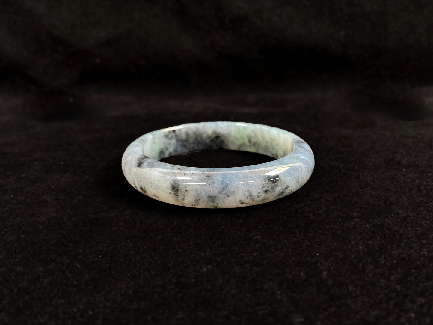 This jadeite bangle features a semi-transparent light green base with evenly distributed charcoal grey inclusions.