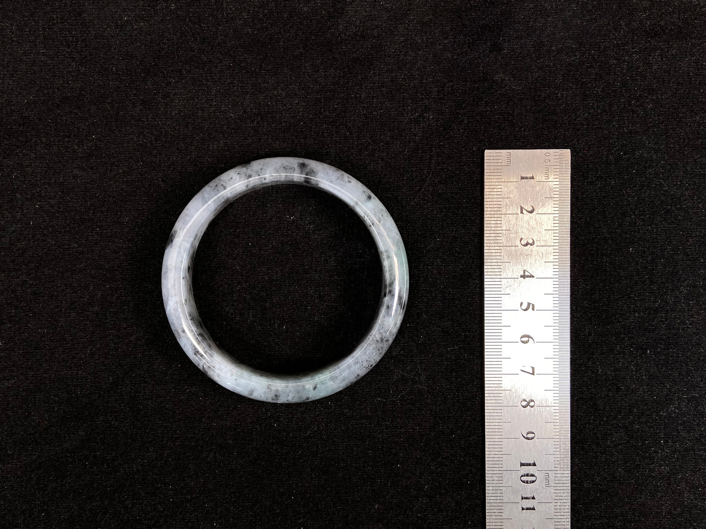 This jadeite bangle features a semi-transparent light green base with evenly distributed charcoal grey inclusions.