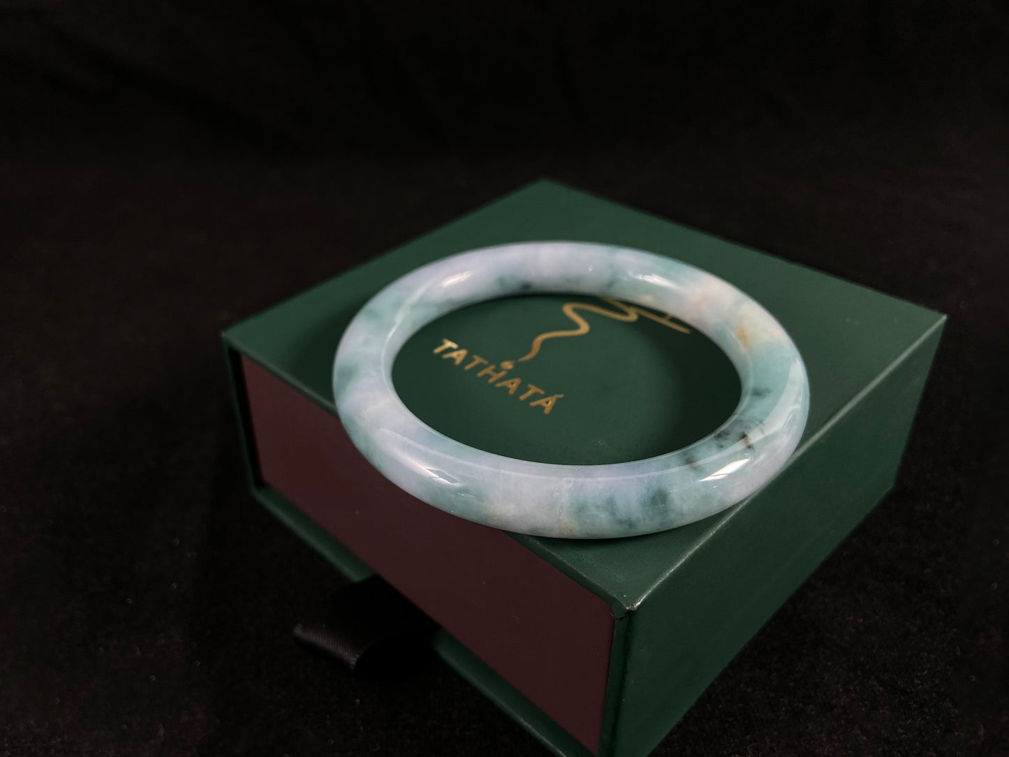 Green jade bracelet on a green box with 'Tathata' branding against a dark background